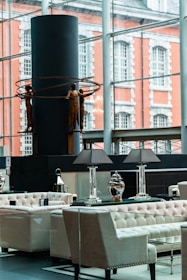 A modern interior space featuring elegant seating arrangements with tufted armchairs and sofas. A table with decorative lamps and vases is prominently placed. In the background, a large window overlooks an exterior with a brick building facade. A contemporary sculpture of human figures holding circular rings is displayed against a dark cylindrical structure.