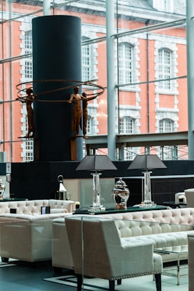 A modern interior space featuring elegant seating arrangements with tufted armchairs and sofas. A table with decorative lamps and vases is prominently placed. In the background, a large window overlooks an exterior with a brick building facade. A contemporary sculpture of human figures holding circular rings is displayed against a dark cylindrical structure.