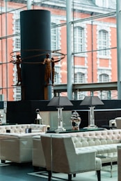A modern interior space featuring elegant seating arrangements with tufted armchairs and sofas. A table with decorative lamps and vases is prominently placed. In the background, a large window overlooks an exterior with a brick building facade. A contemporary sculpture of human figures holding circular rings is displayed against a dark cylindrical structure.