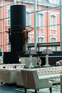 A modern interior space featuring elegant seating arrangements with tufted armchairs and sofas. A table with decorative lamps and vases is prominently placed. In the background, a large window overlooks an exterior with a brick building facade. A contemporary sculpture of human figures holding circular rings is displayed against a dark cylindrical structure.