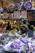 A market stall is filled with vibrant bouquets of flowers wrapped in white paper, predominantly featuring shades of purple and white. A vendor wearing a cap and a mask appears to be arranging the display. Various signs are hanging above the flowers, including payment information and customer instructions. The background shows more floral arrangements and a softly lit market environment.