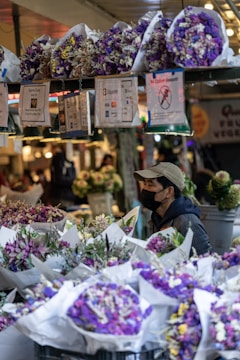 A market stall is filled with vibrant bouquets of flowers wrapped in white paper, predominantly featuring shades of purple and white. A vendor wearing a cap and a mask appears to be arranging the display. Various signs are hanging above the flowers, including payment information and customer instructions. The background shows more floral arrangements and a softly lit market environment.