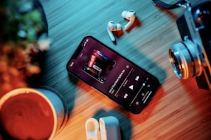 A smartphone on a wooden surface displaying a music player screen with earbuds nearby. The scene includes various items like a potted plant, a camera lens, and a wireless earbud case, creating an organized arrangement. The lighting has a warm and cool contrast, highlighting the textures of the objects.