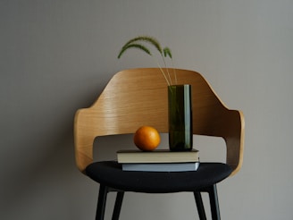 A wooden chair with a sleek, modern design is positioned against a plain background. Resting on the seat is a small stack of books with a bright orange fruit placed on top. Beside the books, a tall, dark green vase holds a few wispy, green plants.