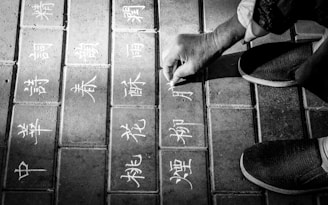 A hand is drawing intricate Chinese characters on square paving stones using chalk. The scene is set on a paved walkway with visible shoe-clad feet indicating someone sitting or kneeling. The overall composition is in black and white, emphasizing the contrast between the chalk and the stones.