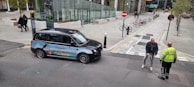 A black taxi with an advertisement on its side is parked beside a road, where two men are having a conversation. The background features a modern urban setting with several buildings, trees, and a bike rack with bicycles. Pavement markings and signs are visible, indicating a bicycle lane and no entry zone.