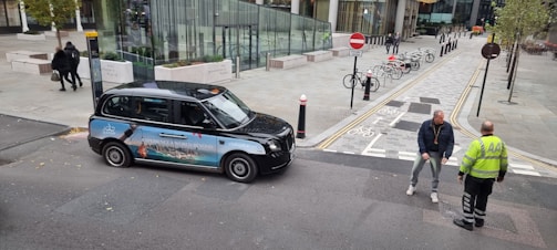 A black taxi with an advertisement on its side is parked beside a road, where two men are having a conversation. The background features a modern urban setting with several buildings, trees, and a bike rack with bicycles. Pavement markings and signs are visible, indicating a bicycle lane and no entry zone.