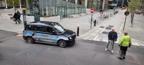 A black taxi with an advertisement on its side is parked beside a road, where two men are having a conversation. The background features a modern urban setting with several buildings, trees, and a bike rack with bicycles. Pavement markings and signs are visible, indicating a bicycle lane and no entry zone.