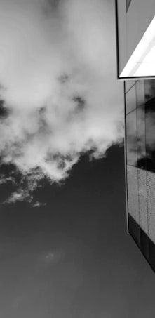 A high-contrast black and white image featuring a modern building with reflective glass panels on the right side. The sky is filled with thick, textured clouds, providing a dramatic backdrop.
