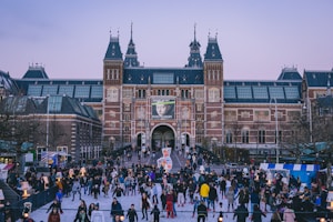 A bustling outdoor ice skating rink is filled with people enjoying the activity in front of an impressive historical building with ornate architecture and spires. The atmosphere is lively with skaters and onlookers around the rink. Banners and decorations add to the festive environment.