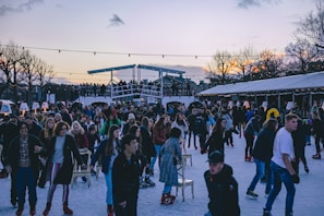 A vibrant scene of people enjoying ice skating on an outdoor rink with a lively crowd gathered around. The setting is festive with string lights overhead and trees in the background, hinting at a winter or holiday season. Some people are skating while others are standing or sitting on chairs placed on the ice. The sky is gradually darkening, with hues of orange and purple visible at the horizon.