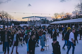 A vibrant scene of people enjoying ice skating on an outdoor rink with a lively crowd gathered around. The setting is festive with string lights overhead and trees in the background, hinting at a winter or holiday season. Some people are skating while others are standing or sitting on chairs placed on the ice. The sky is gradually darkening, with hues of orange and purple visible at the horizon.
