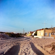 Sandy beach with visible tire tracks leading to a small building labeled 'Policia Maritima'. A yellow boat is stationed nearby, with a clear blue sky overhead and some greenery around.