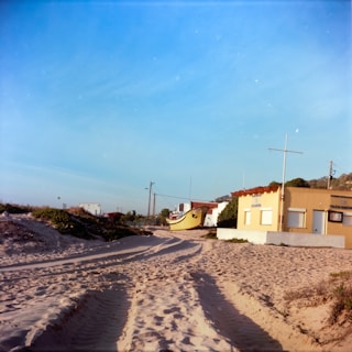 Sandy beach with visible tire tracks leading to a small building labeled 'Policia Maritima'. A yellow boat is stationed nearby, with a clear blue sky overhead and some greenery around.
