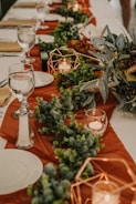 A beautifully set table with a white cloth and an orange runner. The table is adorned with lush green foliage arrangements and copper geometric candle holders containing lit candles. Elegant glassware, plates, and beige napkins are neatly arranged, creating an inviting and sophisticated dining setup.