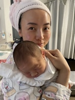 A woman with a towel wrapped around her head is cradling a newborn baby in her arms. The baby is asleep, dressed in a white outfit, and resting peacefully against her shoulder. The setting appears to be indoors, possibly a hospital or home environment with neutral-colored curtains in the background.