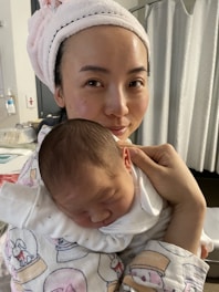 A woman with a towel wrapped around her head is cradling a newborn baby in her arms. The baby is asleep, dressed in a white outfit, and resting peacefully against her shoulder. The setting appears to be indoors, possibly a hospital or home environment with neutral-colored curtains in the background.