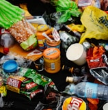 An assortment of discarded food packaging, including plastic bags, bottles, jars, and snack wrappers, strewn across a dark surface. Items such as a loaf of bread, jars labeled Tostitos, a plastic bottle of water, and various snack wrappers are visible.