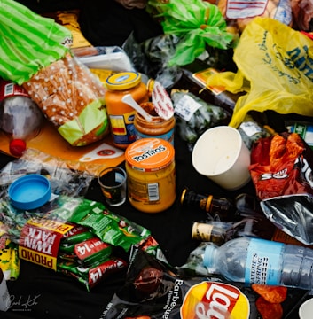 An assortment of discarded food packaging, including plastic bags, bottles, jars, and snack wrappers, strewn across a dark surface. Items such as a loaf of bread, jars labeled Tostitos, a plastic bottle of water, and various snack wrappers are visible.