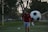 A man in sports attire stands on a soccer field, near a goalpost. He is wearing a red long-sleeve shirt and gray shorts, with a focused or pensive expression. Behind him, a large soccer ball is leaning against the net of the goal.