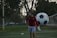 A man in sports attire stands on a soccer field, near a goalpost. He is wearing a red long-sleeve shirt and gray shorts, with a focused or pensive expression. Behind him, a large soccer ball is leaning against the net of the goal.