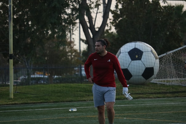 A man in sports attire stands on a soccer field, near a goalpost. He is wearing a red long-sleeve shirt and gray shorts, with a focused or pensive expression. Behind him, a large soccer ball is leaning against the net of the goal.