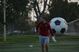 A man in sports attire stands on a soccer field, near a goalpost. He is wearing a red long-sleeve shirt and gray shorts, with a focused or pensive expression. Behind him, a large soccer ball is leaning against the net of the goal.