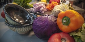 An assortment of fresh vegetables and ingredients is arranged on a kitchen counter. There are ceramic bowls, purple cabbage, orange bell pepper, tomatoes, and sliced red onions, among other items. The scene conveys a sense of preparation for a meal or cooking activity.