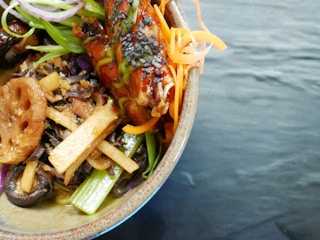 A bowl of colorful Asian-inspired cuisine featuring a variety of fresh vegetables like carrots and scallions, garnished with sesame seeds. The meal includes lotus root, mushrooms, and greens, topped with what appears to be a glazed piece of meat or tofu.