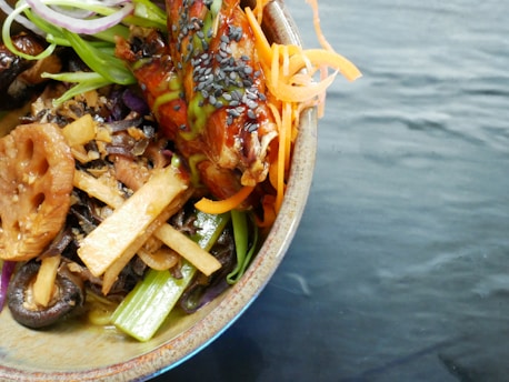 A bowl of colorful Asian-inspired cuisine featuring a variety of fresh vegetables like carrots and scallions, garnished with sesame seeds. The meal includes lotus root, mushrooms, and greens, topped with what appears to be a glazed piece of meat or tofu.