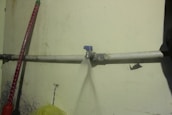 A water pipe is mounted on a wall, with a leak spraying water out in a narrow stream. The wall has some visible stains and damage, and there is a red object leaning against the wall.