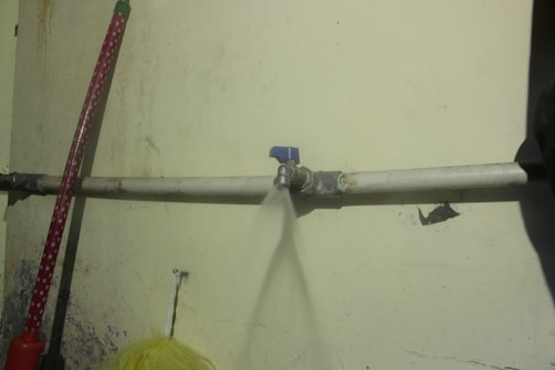 A water pipe is mounted on a wall, with a leak spraying water out in a narrow stream. The wall has some visible stains and damage, and there is a red object leaning against the wall.