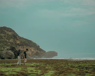 A candid shot of a couple walking hand in hand along a charcoal-colored rocky shoreline.
