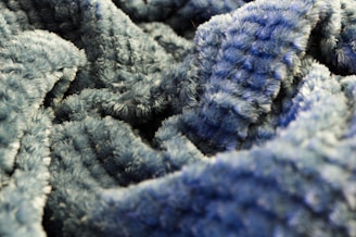 A close-up view of a textured, fluffy blue fabric with a wavy pattern. The fabric appears soft and plush, with a deep pile that suggests warmth and comfort.