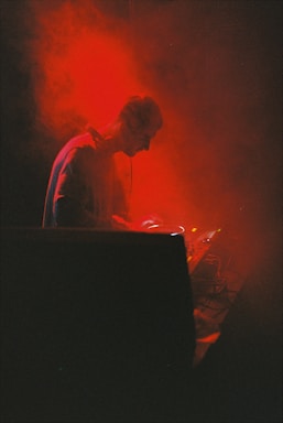 A DJ is deeply focused on mixing music, surrounded by vibrant red smoke and dim lighting, creating an intense and immersive atmosphere.