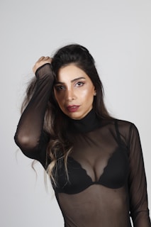 A woman wears a sheer black top over a black bra, with long hair down and makeup accentuating her eyes and lips. She poses with one hand holding her hair against a simple gray background.