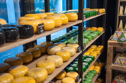 A store shelf displays a variety of cheese wheels in different colors and sizes, organized neatly on wooden shelves. The cheeses range from black and yellow to green, indicating various types and flavors. Small blackboards with labels and prices are placed in front of some cheeses. On an adjacent shelf, there are cheese packages shaped like wedges.