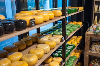 A store shelf displays a variety of cheese wheels in different colors and sizes, organized neatly on wooden shelves. The cheeses range from black and yellow to green, indicating various types and flavors. Small blackboards with labels and prices are placed in front of some cheeses. On an adjacent shelf, there are cheese packages shaped like wedges.