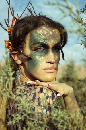A person with elaborate face paint and a nature-inspired headpiece poses among foliage. The face paint features intricate blue and gold designs, adding to an ethereal and artistic appearance. The person wears a detailed, net-like garment and large earrings, complementing the natural aesthetic with delicate blend of colors and textures.