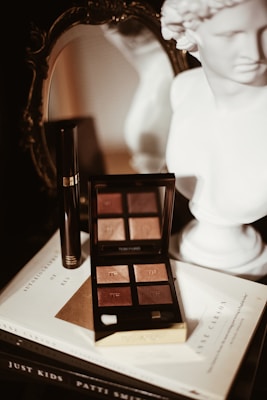 A makeup palette with four shades is placed on top of a stack of books alongside a tube of mascara. A white marble bust and a decorative mirror are in the background.