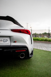 A sleek white sports car is parked on a wet, green grass surface. The car features a prominent rear spoiler, red taillights, and a visible exhaust pipe. A license plate with letters 'GP' is visible. The background includes a gray overcast sky and some distant vegetation and fencing.