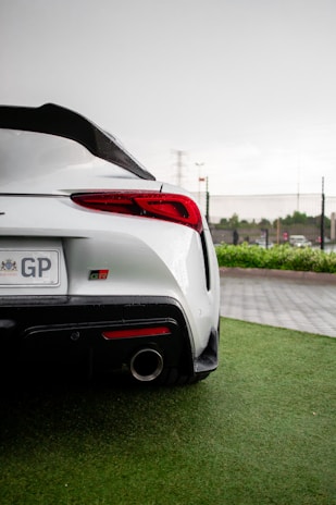 A sleek white sports car is parked on a wet, green grass surface. The car features a prominent rear spoiler, red taillights, and a visible exhaust pipe. A license plate with letters 'GP' is visible. The background includes a gray overcast sky and some distant vegetation and fencing.