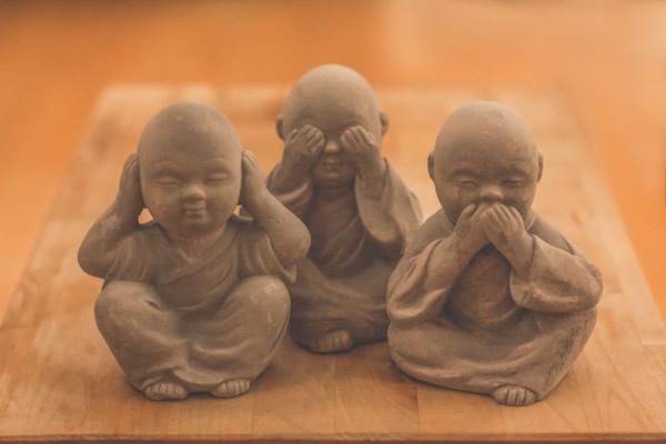 Three small monk statues are sitting in a row on a wooden surface. Each monk has a different gesture: covering their ears, eyes, and mouth. They are made of stone and have a calm and serene expression.
