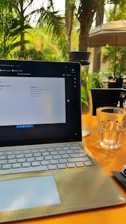 A laptop with a gray keyboard is placed on a bright yellow table. The screen displays a document with a list of items. Next to the laptop, there is a glass of water filled halfway and a tall glass bottle filled with water and mint leaves. The background features lush green palm trees and outdoor seating with black chairs and a large patio umbrella.