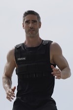 A muscular man is seen wearing a weighted vest, possibly for exercise or training. He has short hair and facial stubble, and appears focused and determined.