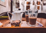 A tray with a white teapot, two glasses filled with tea, and a package of Parle-G biscuits placed on a wooden table in a cozy indoor setting. The background has a white wall adorned with several framed pictures and artworks.