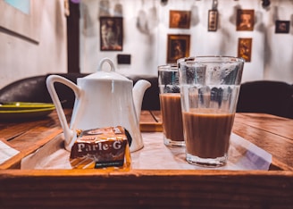 A tray with a white teapot, two glasses filled with tea, and a package of Parle-G biscuits placed on a wooden table in a cozy indoor setting. The background has a white wall adorned with several framed pictures and artworks.