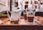 A tray with a white teapot, two glasses filled with tea, and a package of Parle-G biscuits placed on a wooden table in a cozy indoor setting. The background has a white wall adorned with several framed pictures and artworks.