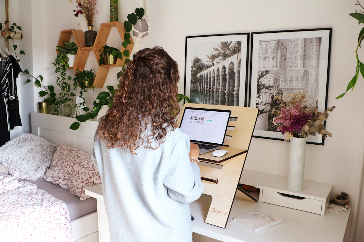 Woman working at standing desk in small plant-filled apartment bedroom with wall shelves and books