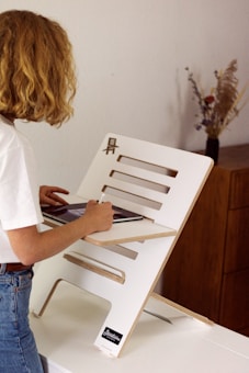 A person with shoulder-length curly hair is working at a standing desk. The desk is made of light-colored wood with grooves and slots, and it supports a tablet or a small laptop. The person is casually dressed in a white t-shirt and jeans. In the background, there is a wooden cabinet with a vase containing dried flowers.